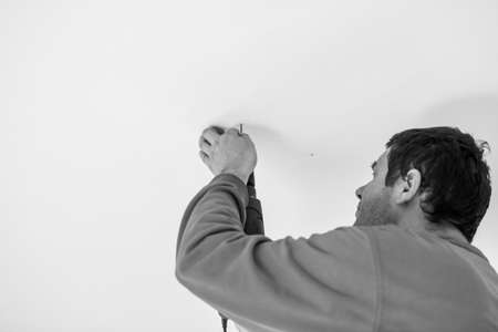 Greyscale image of a workman using a handheld electric drill to drill a hole in the ceiling of a building in a close up view of his head and hands.の写真素材