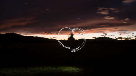 Silhouette of a man draws a heart shape with a flashlight through the air against a colorful orange sunset sky.の写真素材