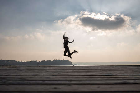 Happy woman jumping in air against sky with sun behind cloud.