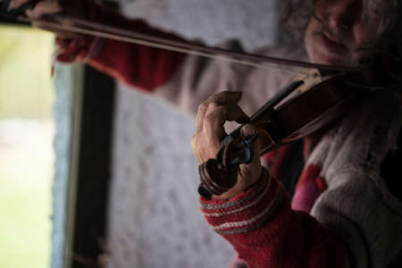 Old man in red sweater full of holes with dirty nails playing a classical violin at home by the window.の写真素材