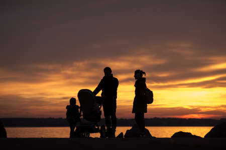 Father and mother with a baby in a stroller and children silhouetted against a colorful orange sunset .の写真素材