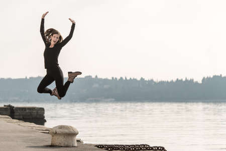 Young woman jumping for joy above a bollard on a quay overlooking a lake or the ocean on a misty atmospheric day.の写真素材