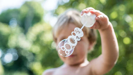 Front view of a young blond child blowing soap bubbles while standing outdoors in nature.
