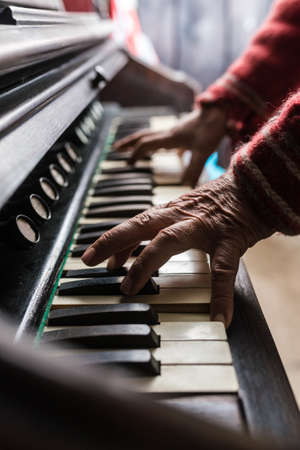 Elderly man playing the organ in a close up view of his wrinkled hands on the keyboard indoors at...