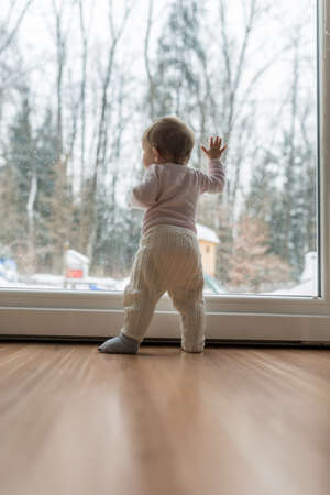 Toddler girl standing up against dirty window looking out to observe nature and forest.の写真素材