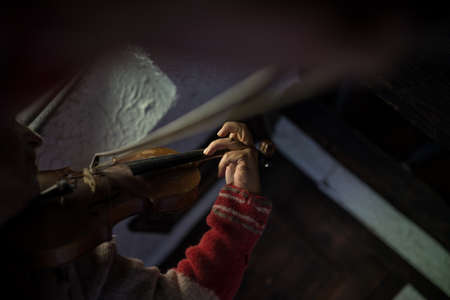 Man in a tattered old torn jersey playing a violin in a darkened room in a beam of light.の写真素材