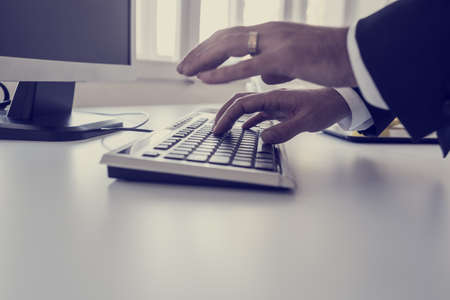 Businessman typing on a computer keyboard on a white desk or office table with foreground copy space in a close up view of his hands, retro toned image.の写真素材