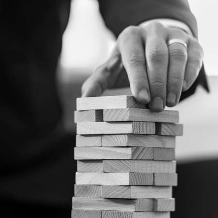 Black and white square format image of businessman placing wooden domino in a tower.の写真素材