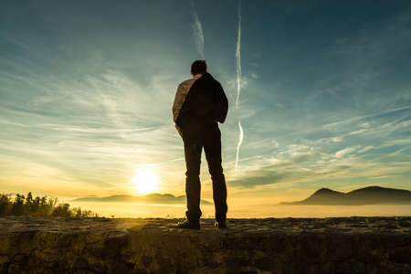 Businessman in a suit standing outdoors silhouetted against a colorful sunrise with the fiery sun peeping over distant mountains with copy space.の写真素材