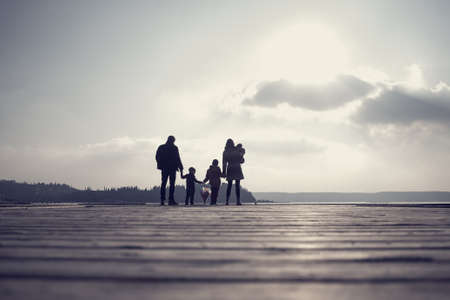 Retro toned image of family with mother, father, two kids and a baby holding hands on a wooden deck overlooking the ocean or lake at sunset.の写真素材