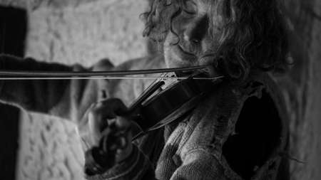Man with long hair and beard in old torn sweater playing a violin, black and white image.