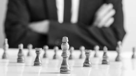 Monochrome image of man wearing suit sitting with folded arms in front of dark chess pieces at white table.の写真素材