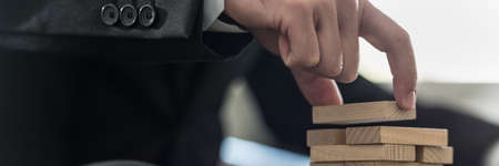 Wide panorama view of businessman placing wooden domino in a tower.