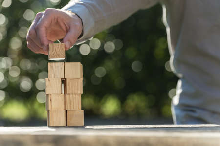 Man building a tower of blank wooden blocks outdoors in the garden in a close up view.の写真素材