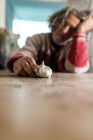 Man with dirty hands holding a bulb of garlic as he sits at table resting his head on his hand in conceptual image of poverty.の写真素材