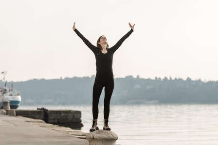 Young brunette standing on bollard with her arms raised high above her head looking up in the sky...
