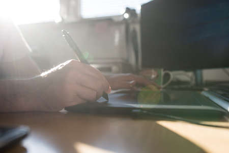 Graphic designer or illustrator using a tablet and stylus pen to do processing and drawing with a bright sunburst through the window pane.の写真素材