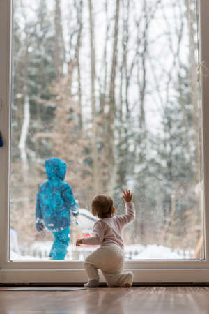 Little baby kneeling in front of a large view window watching a sibling playing in snow in a rural...