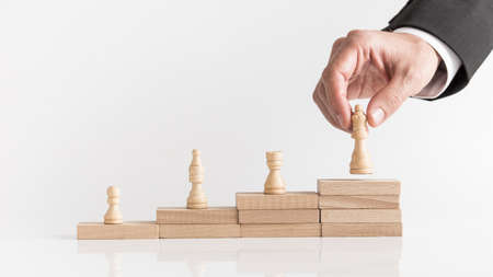 Four chess pieces on a stepped stack of wooden blocks with a businessman placing a queen piece on...