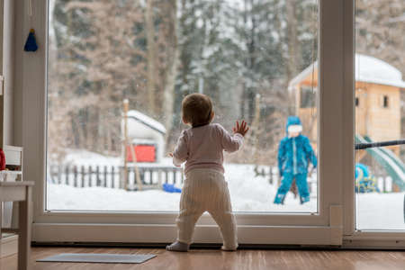 Little baby standing at a large view window watching a sibling outdoors playing in thick winter snow in the garden with forest backdrop.の写真素材