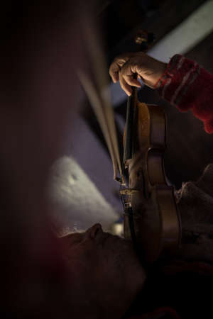 Side bottom view of man with gray beard playing a violin in a shadowy room with focus to the strings and bow.の写真素材