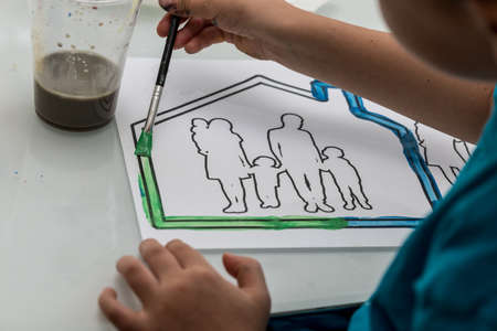 Young child colouring in a sketch of a family in a house using a paintbrush and watercolour paints in an over the shoulder view of the artwork.の写真素材