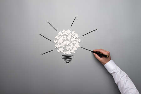 White scattered puzzle pieces and male hand drawing light bulb on grey background.の写真素材