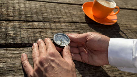 Hands of a man holding compass next to orange coffee cup over old rustic wooden table.