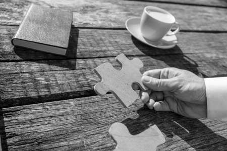 Greyscale close up on hand of unidentifiable business man holding puzzle piece over rustic wooden table for concept about solutions to problems.の写真素材