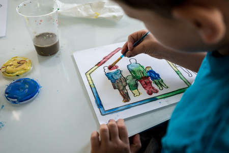 Young boy coloring in an outline sketch of a family in a house with paint carefully filling in the silhouettes with bright colours.の写真素材