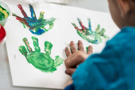 Creative little boy painting with his hands making colorful palm prints on a sheet of paper in an over the shoulder view of the art.の写真素材