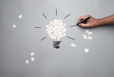 Conceptual image of business vision and idea with a pile of white scattered puzzle pieces forming a light bulb and male hand drawing rays of light on a grey background.の写真素材