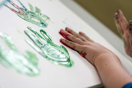 Little child having fun with finger paints creating colourful palm prints on a sheet of white paper at kindergarten.の写真素材