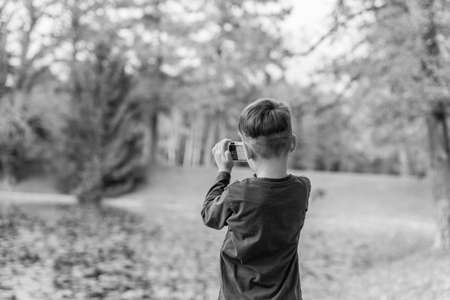 Black and white image of a little boy photographing nature with a compact camera outdoors.
