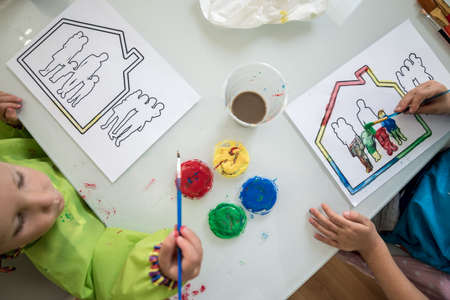 Two children painting a sketch of a family in a house with colourful acrylic paints, top view.の写真素材