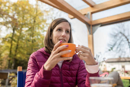 Young woman drinking coffee in orange cup sitting outdoors in autumn day wearing purple jacket.
