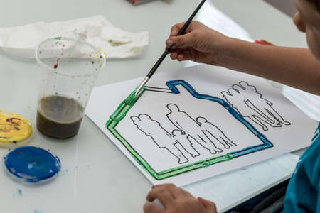 Closeup of toddler boy painting a house with family silhouette inside printed on a white paper.