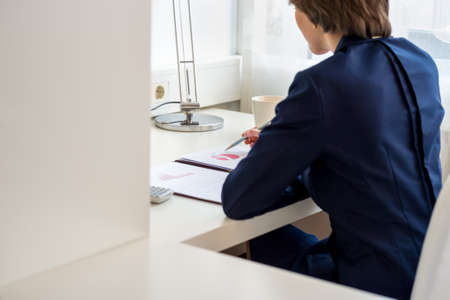 View from behind of a young businesswoman sitting at her desk reviewing financial report with graphical pie chart.の写真素材