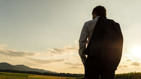 View from behind of a businessman standing with jacket over his shoulder looking at beautiful nature of corn fields and meadows.