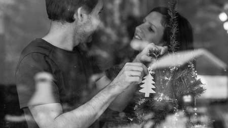 Greyscale image of a loving young couple decorating Christmas tree indoors. View through a window...