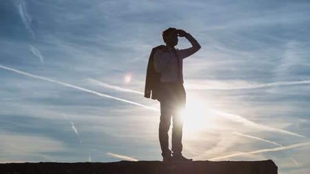 Businessman in a suit standing on a wall saluting the setting sun silhouetted against the sky with...