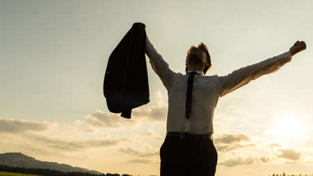 Businessman standing in evening nature with his arms raised up high in the air holding his business jacket in one hand as he celebrates success.の写真素材
