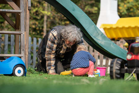 Grandpa playing with his granddaughter outside in backyard as they sit on the grass under a slide and play with sand toys and buckets.の写真素材