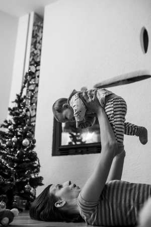 Greyscale image of happy young mother lying on the floor lifting her baby girl in the air as they enjoy Christmas holidays.の写真素材