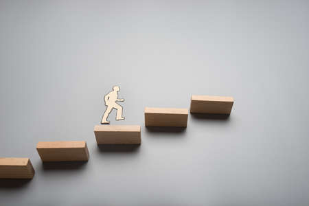 Paper cutout of a businessman walking up the ladder of success made of wooden blocks over grey background.の写真素材