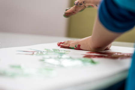 Closeup low angle view of toddler child making colourfull palm prints with finger paints on a piece of white paper.の写真素材