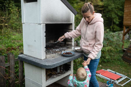 Young mother roasting chestnuts outside in backyard and handing one of them to her toddler daughter.の写真素材