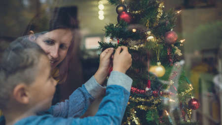 Mother and son decorating Christmas tree together. View through a window.の写真素材