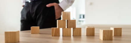 Wooden cubes stacked in pyramid on office desk with businessman standing in background.の写真素材
