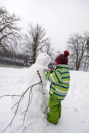 Boy wearing knitted cap making a winter snowman in a snowy landscape.の写真素材